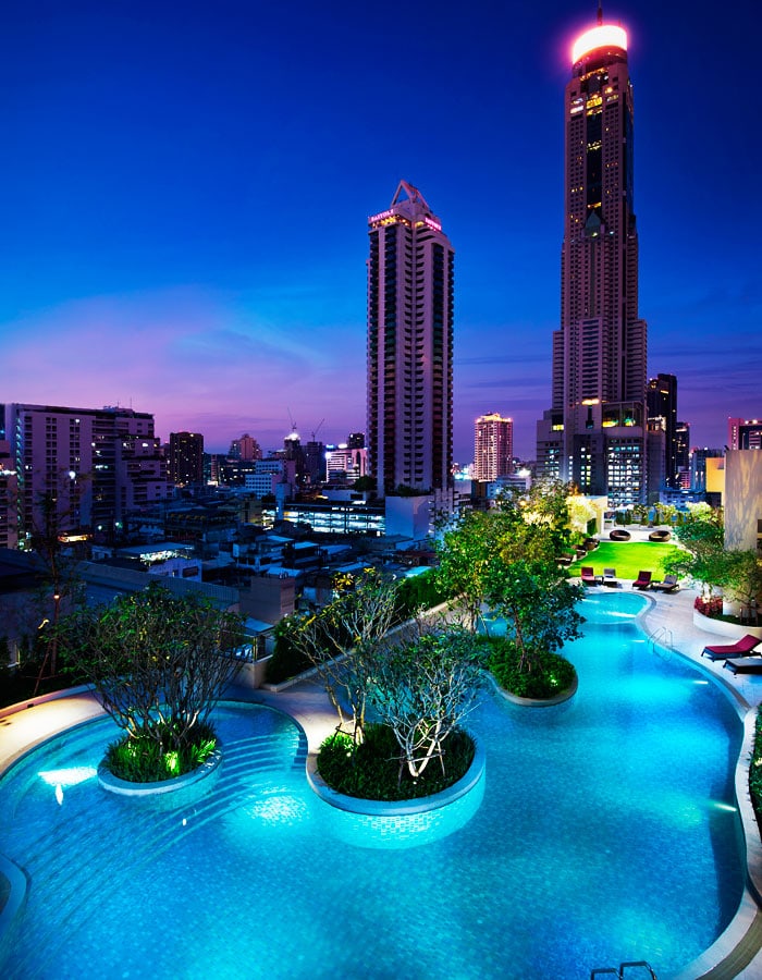 A rooftop pool glows with blue lights at dusk, surrounded by trees and lounge chairs, with a city skyline and tall illuminated skyscrapers in the background.