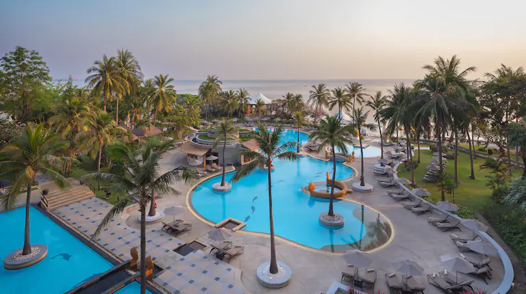 A scenic view of a tropical resort with palm trees, multiple swimming pools, lounge chairs, and pathways, overlooking a calm ocean at sunset or sunrise.