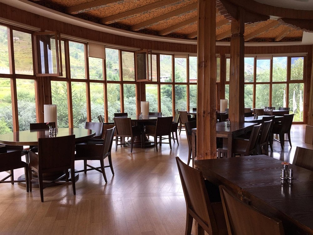 A spacious restaurant with wooden floors, tables, and chairs, featuring large floor-to-ceiling windows that offer scenic views of green hills and trees outside. Natural light fills the room.