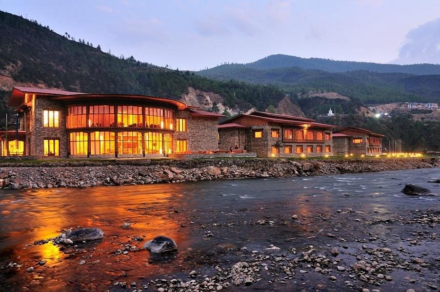 A modern riverside building with large windows glows warmly at dusk, reflecting on the water, surrounded by forested hills and mountains under a soft evening sky.
