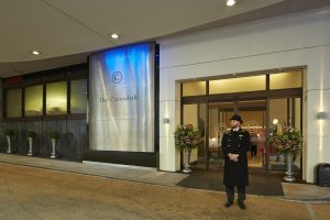 A doorman in uniform stands outside the entrance of The Cavendish hotel, which features large glass doors, flower arrangements in vases, and a modern sign with the hotel name on the exterior wall.