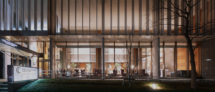 Exterior view of a modern building at night with large glass windows revealing a brightly lit interior. A sign in front reads “THE CELESTINE.” Trees and grass are visible in the foreground.