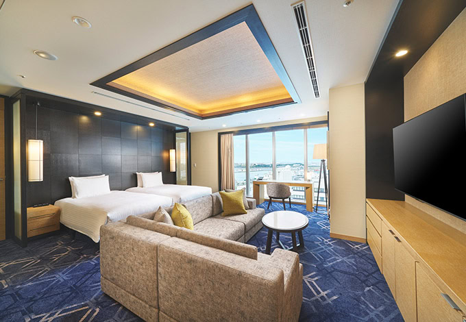 A modern hotel room features two beds, a beige sofa, a round table with chairs, a large flat-screen TV, and floor-to-ceiling windows offering a city view. The room has elegant lighting and blue-patterned carpet.