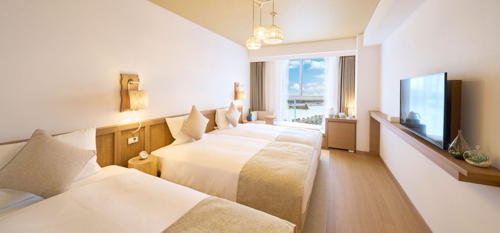 A bright, modern hotel room with three large beds, neutral bedding, wood accents, wall lamps, a flat-screen TV, and a large window offering a scenic view of water and sky.