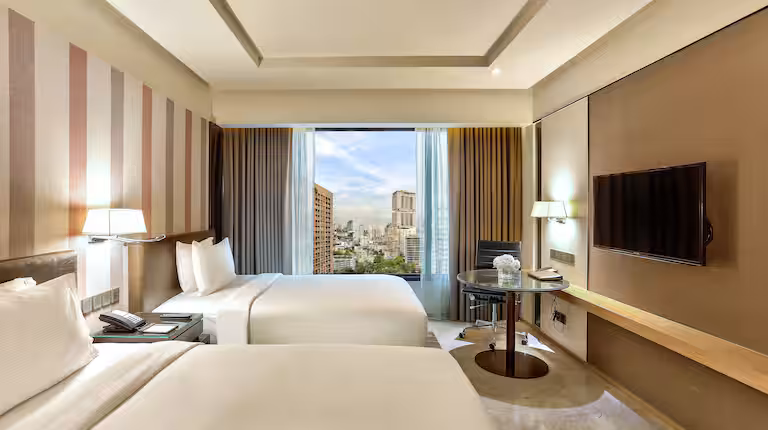 A modern hotel room with two double beds, striped accent wall, wall-mounted TV, desk with chair, and large window offering a city view with tall buildings under a partly cloudy sky.