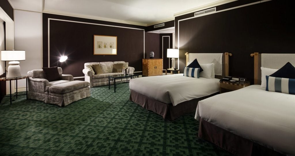 A hotel room with two double beds, a green patterned carpet, a beige sofa and armchair, bedside tables with lamps, a small table, and dark brown walls with white trim.