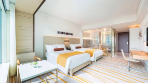 A modern hotel room with two double beds, each with white linens and yellow throws. The room features large windows, a desk, chairs, a small table, and a glass-walled bathroom in the background.
