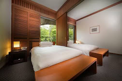 A modern hotel room with two single beds, wooden furniture, large windows showing greenery outside, bedside lamps, and a framed picture on the wall.