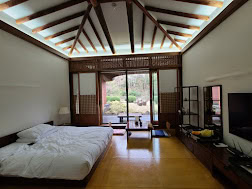 A cozy room with a low bed, wooden beams on the ceiling, sliding doors opening to a garden, and a TV with a fruit basket on a low table; soft natural light fills the space.