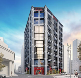 A modern, multi-story black building with large windows and red accents stands on a city corner under a blue sky. The bottom floor has glass entrances, and a few people walk nearby on the street.