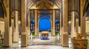 Spacious open-air hotel lobby with elegant arches, wooden ceiling, lanterns, and a view through to the ocean in the distance at dusk.