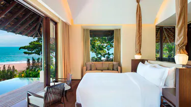 A bright, modern hotel room with a large bed, seating area, and floor-to-ceiling windows offering views of a lush garden, private pool, and the ocean.