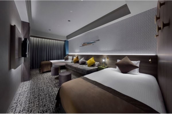 Modern hotel room with two beds, a long sofa, accent pillows, wall-mounted TV, small round tables, patterned carpet, and soft, ambient lighting. Curtains cover a large window at the end of the room.