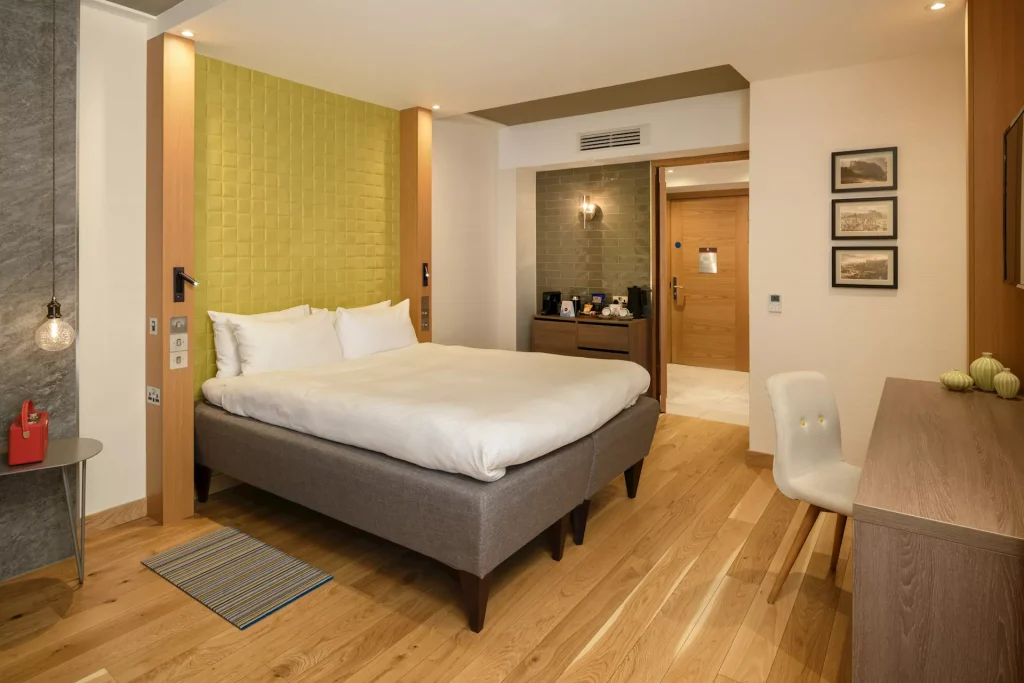 Modern hotel room with a double bed, green accent wall, wooden floor, desk with chair, and a dresser with a coffee station. The room has warm lighting and minimalist decor.