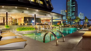 Modern outdoor hotel pool at night with lounge chairs, greenery, and city buildings in the background, illuminated by warm lighting. Steps lead into the clear water, and the pool area appears inviting and tranquil.