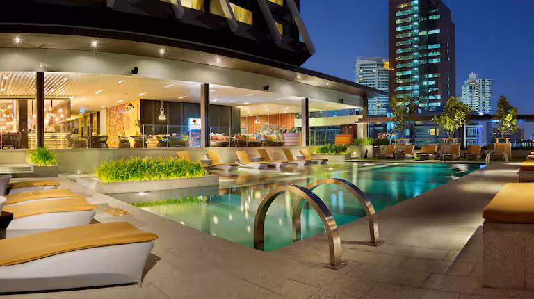 Modern outdoor hotel pool at night with lounge chairs, greenery, and city buildings in the background, illuminated by warm lighting. Steps lead into the clear water, and the pool area appears inviting and tranquil.