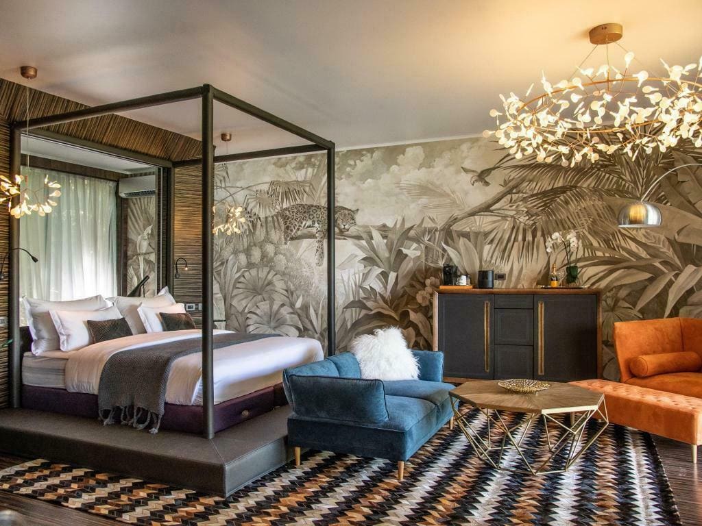 Stylish hotel room with a four-poster bed, velvet blue sofa, orange armchair, geometric rug, gold coffee table, and a jungle-themed mural. Modern lighting fixtures hang from the ceiling.