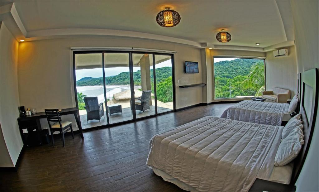 A spacious hotel room with two beds, a desk, and a TV, featuring large glass doors that open to a balcony with chairs and a view of a beach and lush green hills.