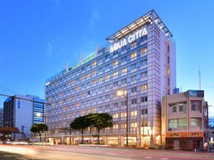 A modern, multi-story hotel named AQUA CITTA is illuminated at dusk, with a glass exterior and rooftop lights. Cars pass in front, and other buildings are visible nearby under a clear evening sky.