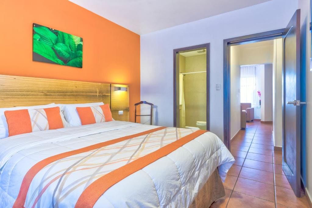 A brightly lit hotel room with a bed covered in white and orange linens, matching pillows, and a wooden headboard. An orange accent wall features a green leaf artwork. Two open doors reveal a bathroom and another room.