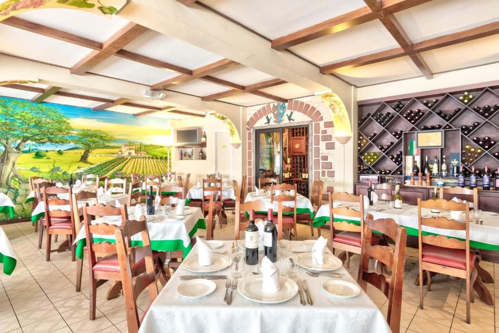 Bright, inviting restaurant with wooden chairs and tables set with white tablecloths and wine bottles. A colorful mural of a vineyard decorates one wall, and a wine rack is visible near the bar area.