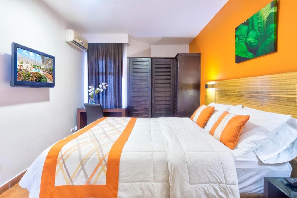 A modern hotel room with a double bed, white and orange bedding, a wooden headboard, and matching orange pillows. There is a desk with a chair, a wardrobe, wall art, and a TV mounted on the wall.