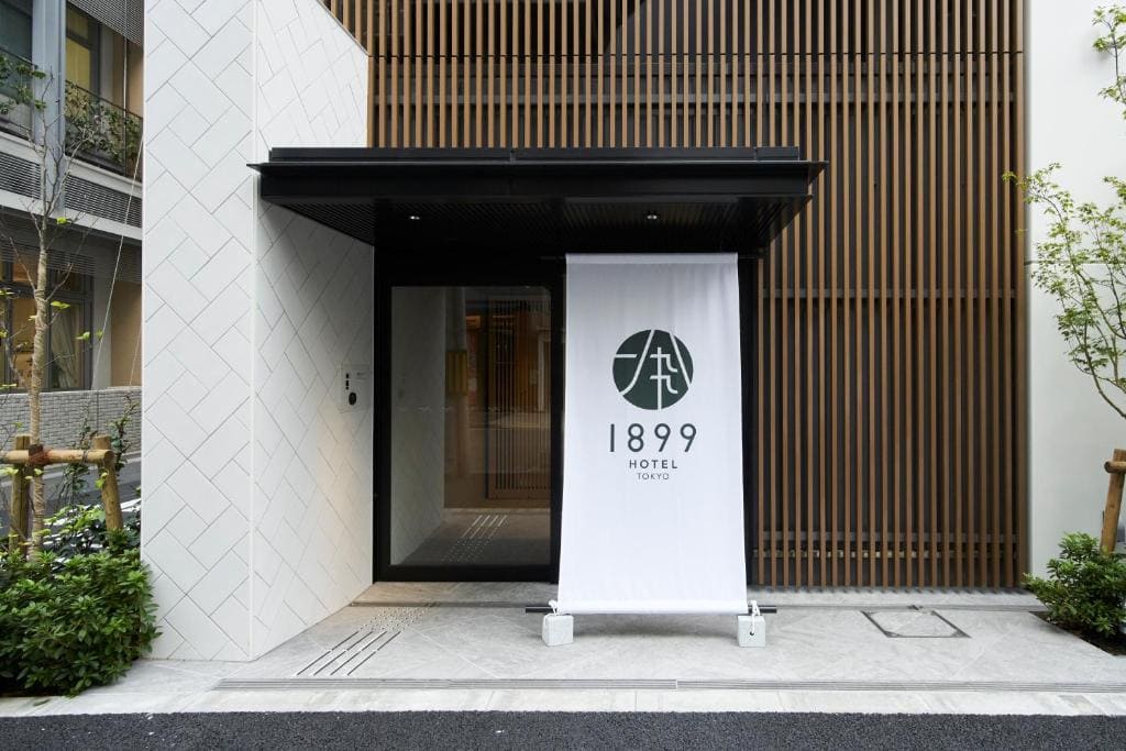 Entrance of a modern hotel with a white banner displaying 1899 HOTEL TOKYO and a green logo, surrounded by plants and wooden slats on the building facade.