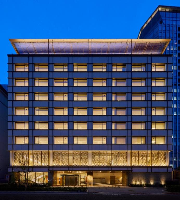 A modern, multi-story hotel building is illuminated with warm yellow lights at dusk, with a flat, overhanging roof and large glass windows evenly spaced across the facade.