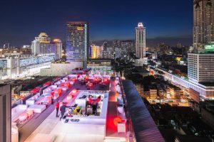 A vibrant rooftop bar with glowing red and white lights overlooks a bustling city skyline at night, surrounded by tall buildings and illuminated streets below.