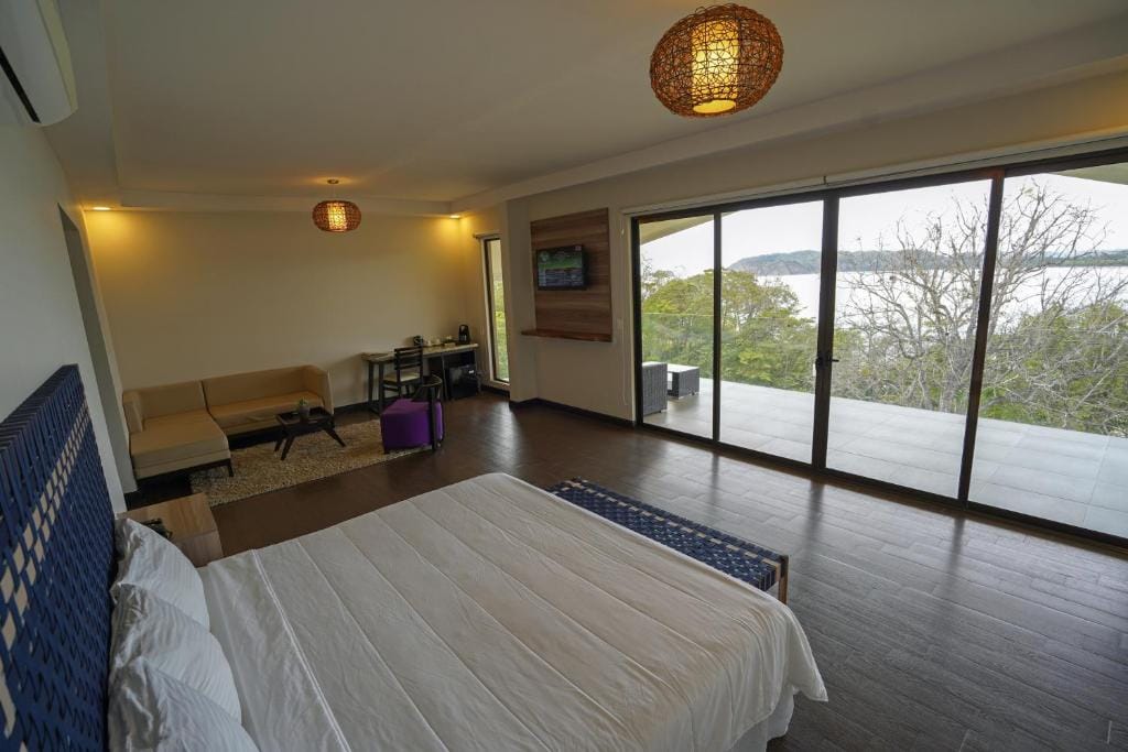 A spacious, modern bedroom with a large bed, a sofa, desk, and sliding glass doors leading to a balcony with scenic views of trees and water. Warm lighting and wooden floors create a cozy atmosphere.