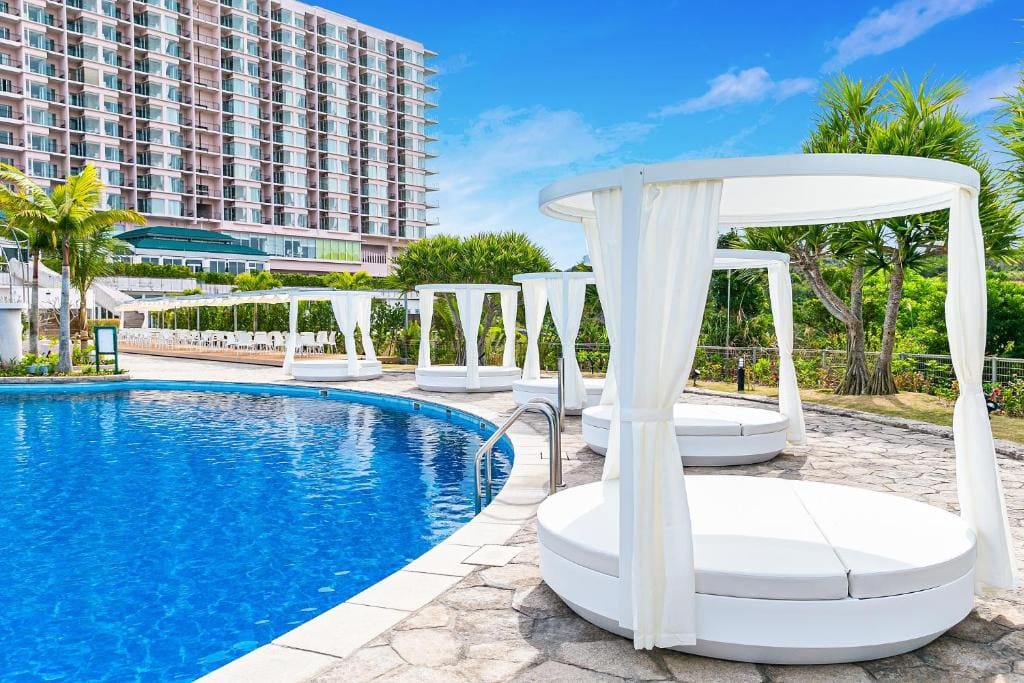 A sunny outdoor pool scene with round white canopy beds along the poolside, surrounded by palm trees and greenery, with a modern multi-story hotel building in the background.