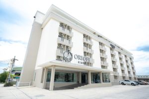 A modern, multi-story hotel building with large windows and balconies, named Oriole Residence, on a bright day. The entrance has a glass facade and the parking lot beside the building is partially visible.