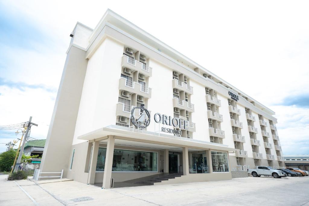 A modern, multi-story hotel building with large windows and balconies, named Oriole Residence, on a bright day. The entrance has a glass facade and the parking lot beside the building is partially visible.