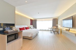 A spacious, modern hotel room with a large bed, red accent pillows, a wall-mounted TV, desk, chair, and a kitchenette area with a microwave and bottled water. Light wood floors and large window with sheer curtains brighten the space.