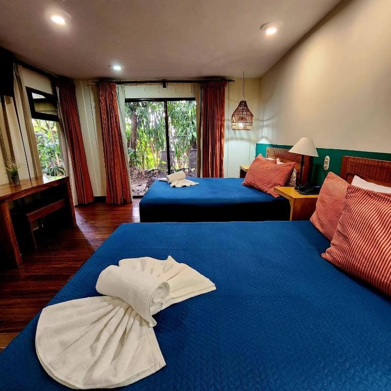 A cozy hotel room with two double beds covered in blue quilts, red pillows, and towels folded in a fan shape. The room has wooden floors, a desk, large windows, and glass doors opening to a lush garden outside.