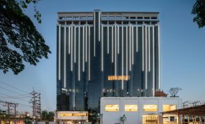 A modern high-rise building with vertical white LED lights on its dark glass facade, standing behind a lower white structure, photographed at dusk with a clear sky and trees framing the scene.