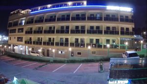 A night view of a multi-story hotel with balconies, well-lit exterior, and several parked cars in front of the building. The parking lot is mostly empty, and blue neon lights highlight the hotel’s roofline.