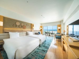 A spacious hotel room with two large beds, light wood furniture, a blue patterned rug, wall-mounted decorations, a desk, a flat-screen TV, and large windows offering a view of the ocean and mountains.