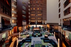 A spacious, modern hotel lobby with high ceilings, multiple floors, lounge seating areas, stylish lighting, patterned rugs, and warm ambient lighting, viewed from an elevated perspective.