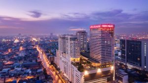 A cityscape at dusk with tall buildings lit up, including a prominent hotel with a red PRINCE PALACE sign. Streets below are illuminated, and the sky is a mix of purple and blue hues.
