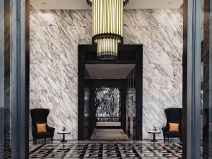 Elegant hotel lobby with marble walls and geometric patterned floor, featuring a large chandelier, two high-back black chairs with orange cushions, and small round tables with flowers on each side of the hallway entrance.