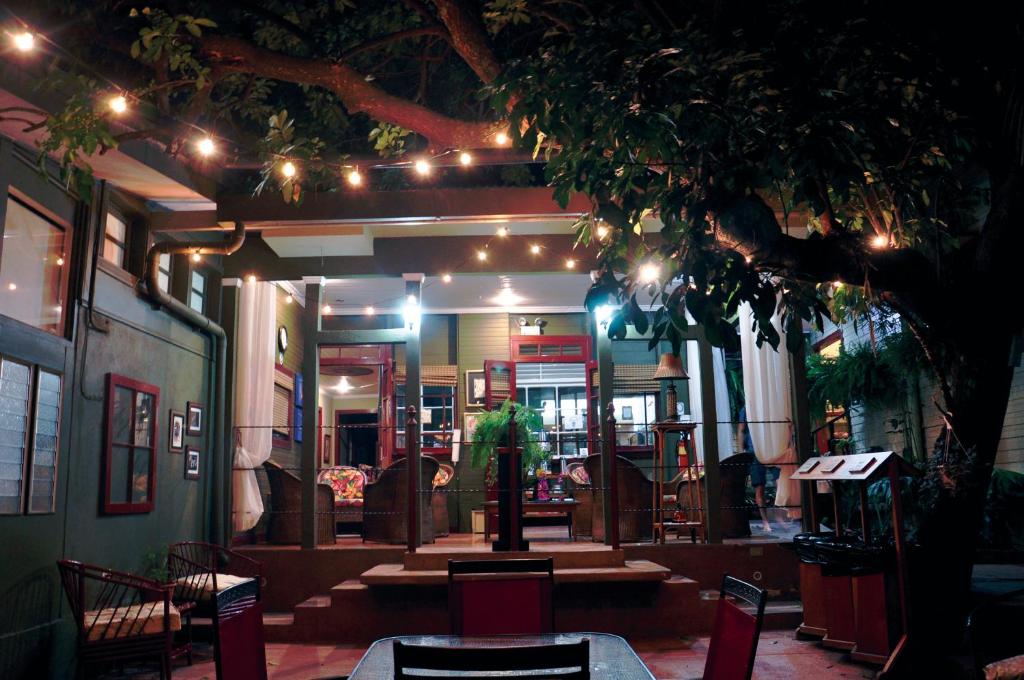 A cozy outdoor patio at night with string lights hanging from a tree, a dining table with red chairs in the foreground, and a warmly lit house with large windows and plants in the background.