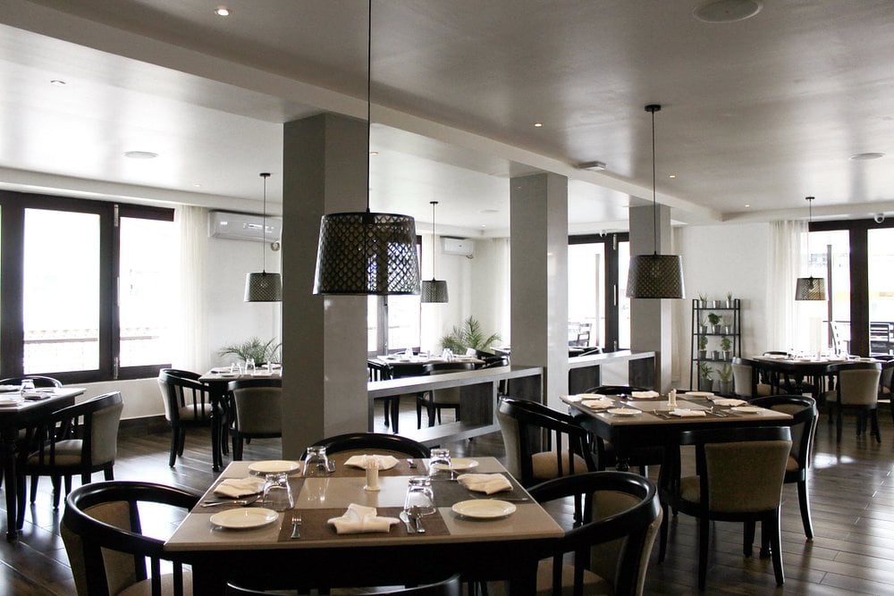 Modern restaurant interior with neatly set square tables, black chairs, pendant lights, large windows, and minimalist decor. The space is bright and inviting with natural light and contemporary furnishings.