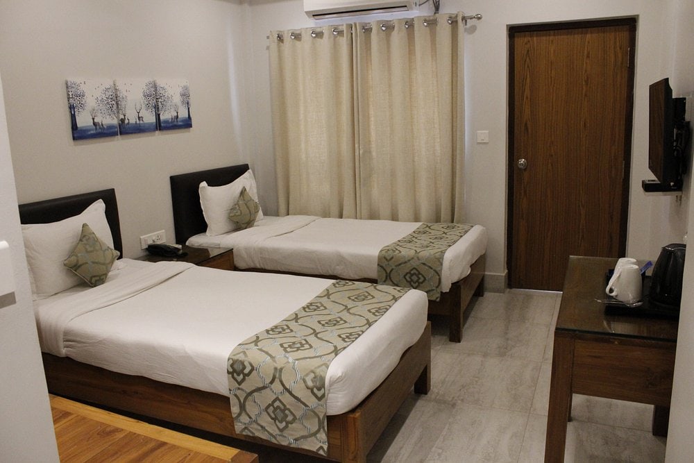 A neatly arranged hotel room with two single beds, white bedding, and decorative runners. There are cream curtains, wall art, a wooden desk with a kettle, and a wall-mounted TV beside a wooden door.