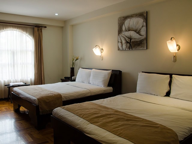 A cozy hotel room with two double beds, beige bedding, a window with sheer curtains, wall-mounted lights, a bedside table with a vase, and a monochrome flower painting above the beds.