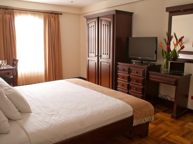 A neatly made bed with white sheets and a tan throw faces a wooden wardrobe, dresser with a TV, and a desk with a mirror and tropical flowers in a vase, in a warmly lit hotel room with wooden flooring.
