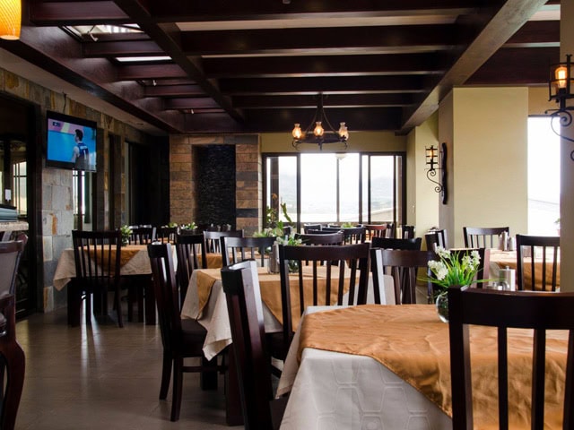 A cozy restaurant with wooden chairs and tables covered in white and tan tablecloths, decorated with small flower arrangements. Large windows let in natural light, and a TV is mounted on the wall.