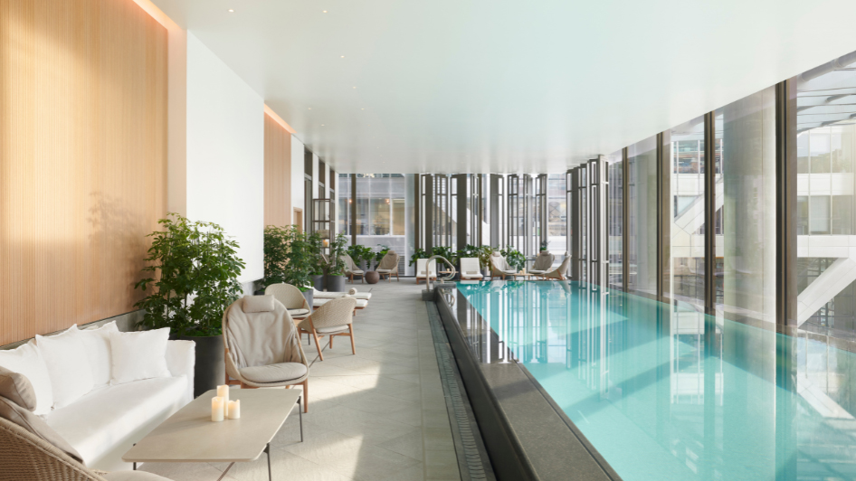 Modern indoor swimming pool area with floor-to-ceiling windows, wooden wall accents, cozy lounge chairs, green plants, and a calm, inviting atmosphere. Natural light fills the elegant, serene space.