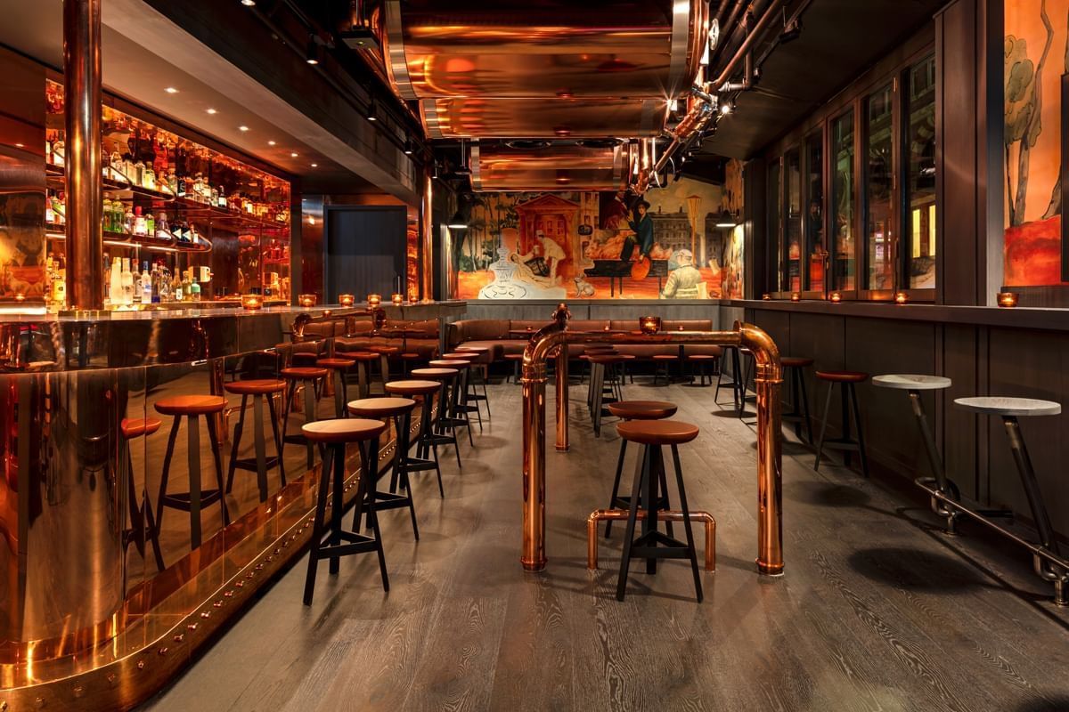 A stylish bar with copper accents, high stools, a reflective bar counter, and colorful wall murals. Warm lighting creates a cozy atmosphere, with small tables and cushioned bench seating along one wall.