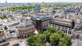 Aerial view of a cityscape with modern and historic buildings, tree-lined streets, and a park with a circular fountain in the foreground, under a bright sky.